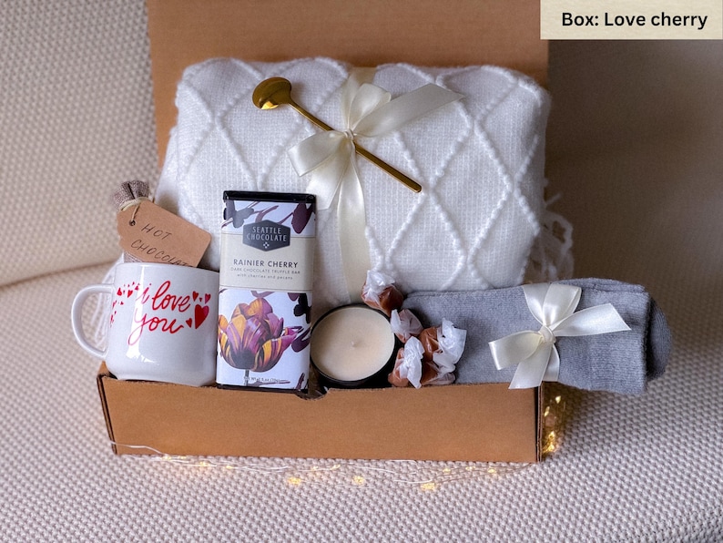 May include: A gift box filled with a white blanket, mug, Seattle Chocolate bar, candle, and gray socks tied with a white ribbon. The mug says "I love you" with red hearts. A gold spoon rests on the blanket.