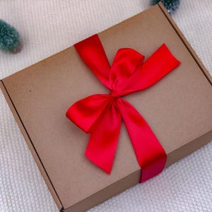 K&ouml;nnte beinhalten: Eine braune Geschenkbox aus Pappe, gebunden mit einem leuchtend roten Satinband und einer Schleife. Die Box ist rechteckig und liegt auf einer wei&szlig;en, strukturierten Oberfl&auml;che. Kleine gr&uuml;ne Deko-B&auml;ume sind um die Box platziert.