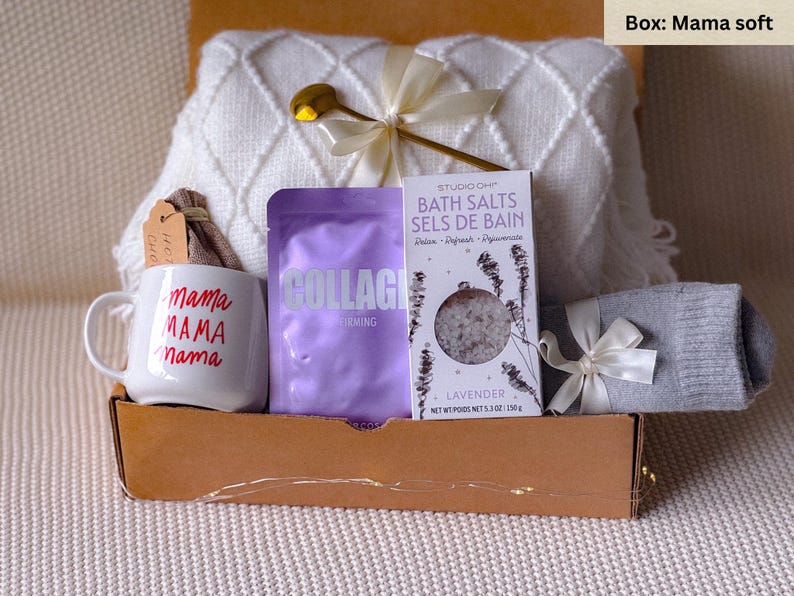 May include: A gift box filled with self-care items, including a white mug with the text "mama MAMA nama" in red, a purple package labeled "COLLAGEN IRMING", a white bath salt package with lavender scent, and a gray knitted sock.