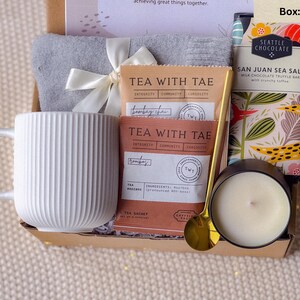 May include: A gift box containing a white ribbed mug, tea sachets, a gray towel tied with a white ribbon, a gold spoon, a candle in a black container, and a chocolate bar. The box is presented on a neutral-colored surface.