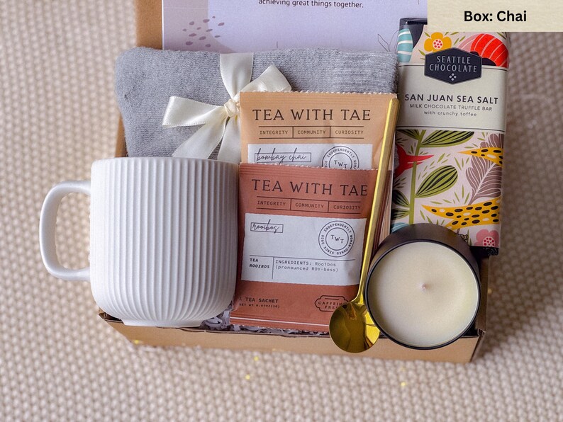 May include: A gift box containing a white ribbed mug, tea sachets, a gray towel tied with a white ribbon, a gold spoon, a candle in a black container, and a chocolate bar. The box is presented on a neutral-colored surface.