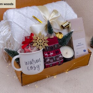 K&ouml;nnte beinhalten: Ein Geschenk-Korb gef&uuml;llt mit einer wei&szlig;en Decke, einer Tasse mit dem Text "warm cozy", roten Socken, einer Schokoladentafel, einer Kerze und dekorativen Gegenst&auml;nden. Der Korb enth&auml;lt kleine Weihnachtsb&auml;ume und einen goldenen Schneeflocken-Ornament.