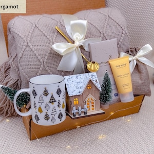 May include: A gift box with a beige blanket, a mug with Christmas tree designs, a wooden house, and a tube of body lotion. The box is tied with a white ribbon and contains a small bag and a golden spoon.