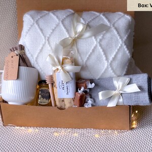 May include: A gift box filled with a white blanket, mug, honey, cookies, socks, and chocolates. The box is brown and tied with white ribbons. The mug has a tag that says "Hot Chocolate".