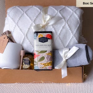 May include: A gift box with a white mug, a jar of honey, a wooden honey dipper, a bar of San Juan Sea Salt chocolate, a white blanket, and a pair of gray socks. The box is tied with white ribbons and includes a tag that says "Hot Chocolate."