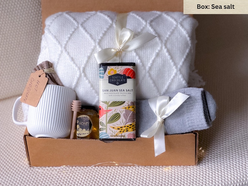 May include: A gift box with a white mug, a jar of honey, a wooden honey dipper, a bar of San Juan Sea Salt chocolate, a white blanket, and a pair of gray socks. The box is tied with white ribbons and includes a tag that says "Hot Chocolate."