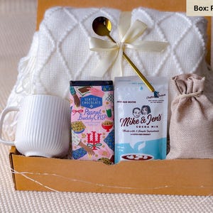 May include: A gift box filled with a white mug, a white textured blanket, a gold spoon, a bag of cocoa mix, a bag of peanut brittle, and a small burlap sack. The box is made of cardboard and tied with a white ribbon.