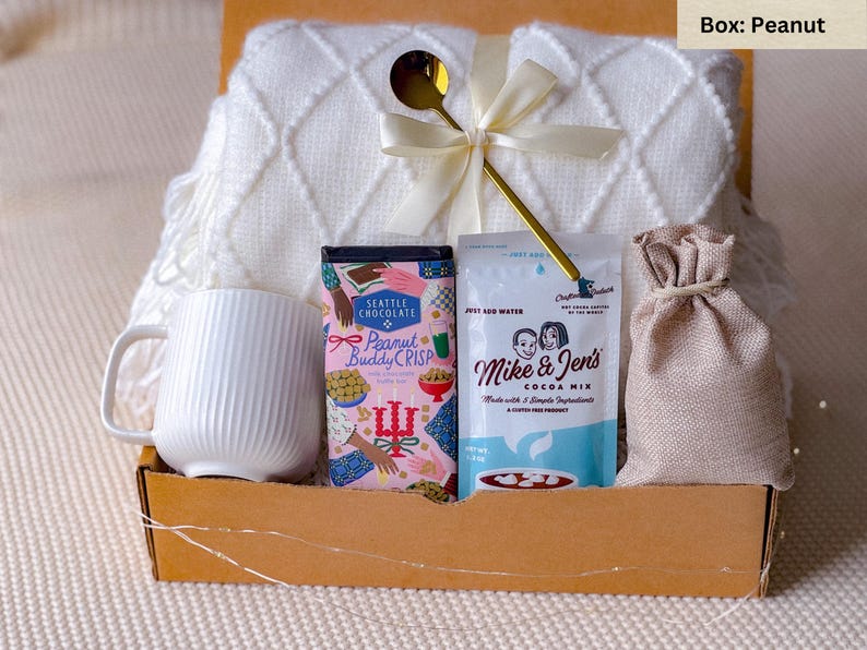 May include: A gift box filled with a white mug, a white textured blanket, a gold spoon, a bag of cocoa mix, a bag of peanut brittle, and a small burlap sack. The box is made of cardboard and tied with a white ribbon.