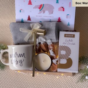 May include: A warm white gift box filled with cozy items, including a mug that says "warm + cozy", a candle, a pair of socks, caramel candies, and a packet of white hot chocolate.