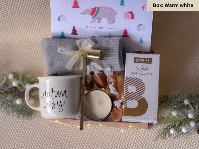 May include: A warm white gift box filled with cozy items, including a mug that says "warm + cozy", a candle, a pair of socks, caramel candies, and a packet of white hot chocolate.