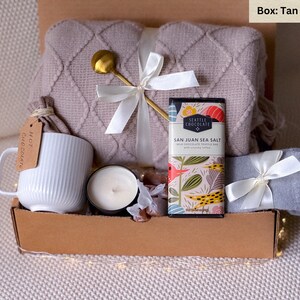 May include: A gift box filled with cozy items, including a tan knitted throw blanket, a white mug with the words "Hot Chocolate" on it, a candle, a chocolate bar with the text "Seattle Chocolate San Juan Sea Salt", and a pair of gray socks.