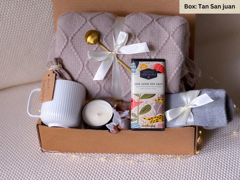 May include: A gift box filled with cozy items, including a tan knitted throw blanket, a white mug with the words "Hot Chocolate" on it, a candle, a chocolate bar with the text "Seattle Chocolate San Juan Sea Salt", and a pair of gray socks.