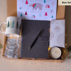 May include: A grey gift box filled with a mug with a winter forest design, a black journal, a package of hot cocoa mix, a candle, and a pen. The box is decorated with a Christmas card featuring a polar bear and the words "Merry Christmas".