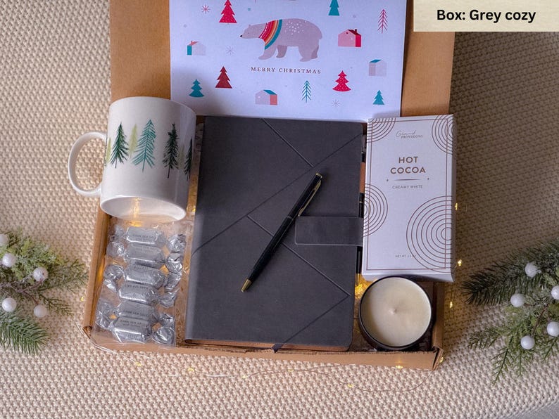 May include: A grey gift box filled with a mug with a winter forest design, a black journal, a package of hot cocoa mix, a candle, and a pen. The box is decorated with a Christmas card featuring a polar bear and the words "Merry Christmas".