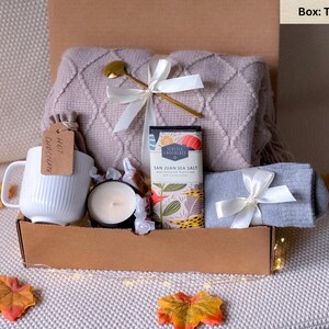 May include: A gift box with a tan salt box containing a white mug, a candle, a chocolate bar, a gray scarf, a blanket, and a gold spoon. The box is decorated with a white ribbon and autumn leaves.