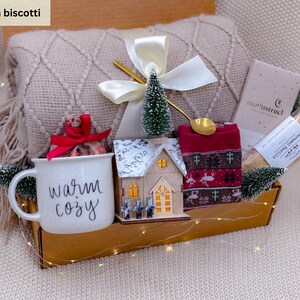K&ouml;nnte beinhalten: Geschenkbox mit beiger Decke, Tasse mit "warm cozy" Schriftzug, Holzhaus, rotem Schal, kleinem Baum und goldenem L&ouml;ffel. Die Box ist mit einer wei&szlig;en Schleife und Lichterketten dekoriert. Enth&auml;lt Kekse.