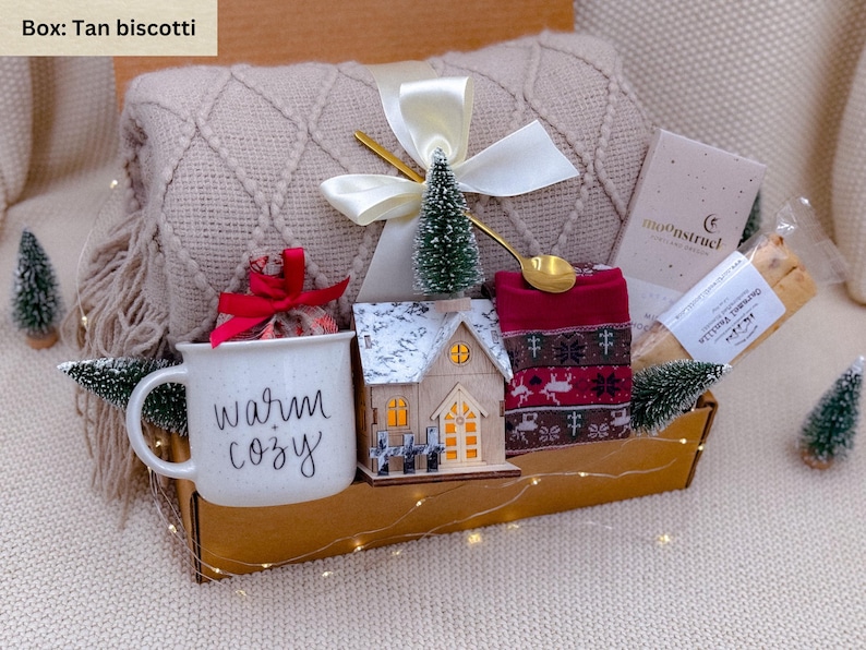 K&ouml;nnte beinhalten: Geschenkbox mit beiger Decke, Tasse mit "warm cozy" Schriftzug, Holzhaus, rotem Schal, kleinem Baum und goldenem L&ouml;ffel. Die Box ist mit einer wei&szlig;en Schleife und Lichterketten dekoriert. Enth&auml;lt Kekse.