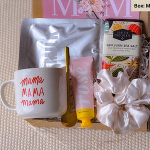 May include: A gift box filled with items for a mother. The box contains a white mug with the text "mama MAMA mama" in red, a gold spoon, a pink hand cream tube with the word "rose" in white, a white satin hair scrunchie, and a chocolate bar with the text "SAN JUAN SEA SALT MILK CHOCOLATE TRUFFLE BAR" and a floral design.