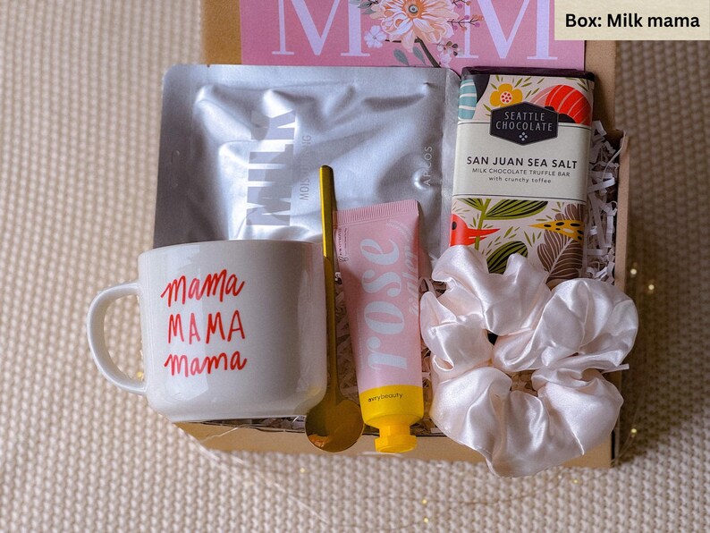 May include: A gift box filled with items for a mother. The box contains a white mug with the text "mama MAMA mama" in red, a gold spoon, a pink hand cream tube with the word "rose" in white, a white satin hair scrunchie, and a chocolate bar with the text "SAN JUAN SEA SALT MILK CHOCOLATE TRUFFLE BAR" and a floral design.