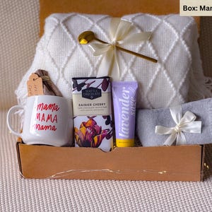 May include: A gift box labeled "Mama dream" contains a white mug with "mama" in red, a white blanket, chocolate, lavender sage lotion, gray socks, and a gold spoon. The box is brown.