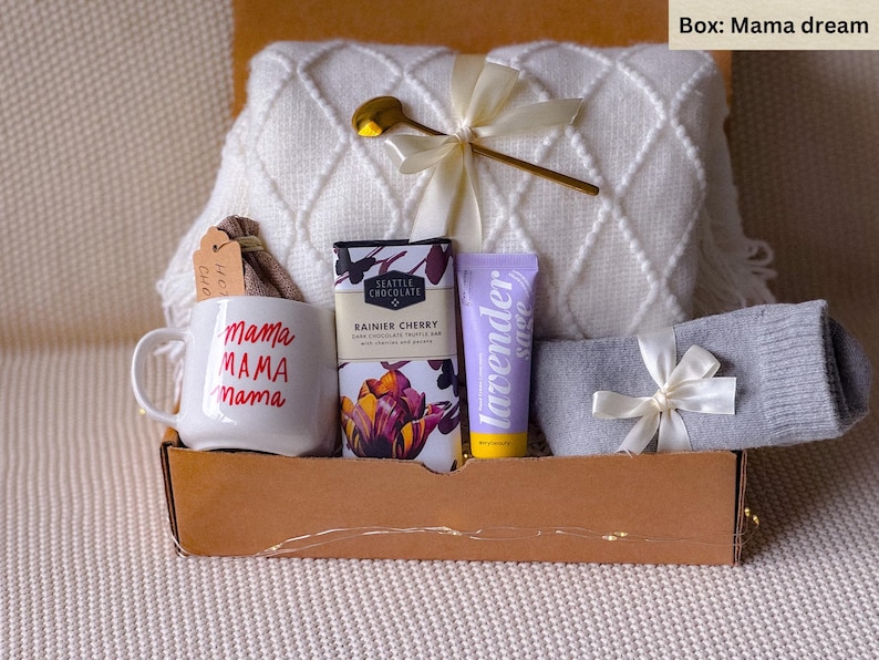 May include: A gift box labeled "Mama dream" contains a white mug with "mama" in red, a white blanket, chocolate, lavender sage lotion, gray socks, and a gold spoon. The box is brown.