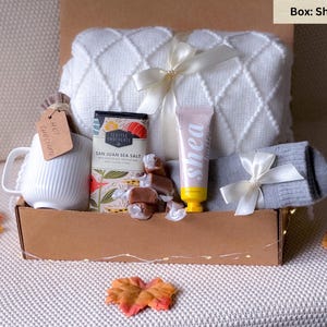 May include: A gift box filled with cozy items for a relaxing evening. It includes a white blanket, a mug, a bar of chocolate, a tube of shea butter, a pair of socks, and caramel candies. The box is labeled "Shea Salt".