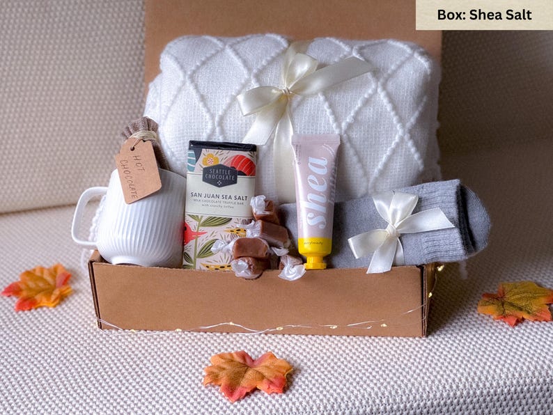 May include: A gift box filled with cozy items for a relaxing evening. It includes a white blanket, a mug, a bar of chocolate, a tube of shea butter, a pair of socks, and caramel candies. The box is labeled "Shea Salt".