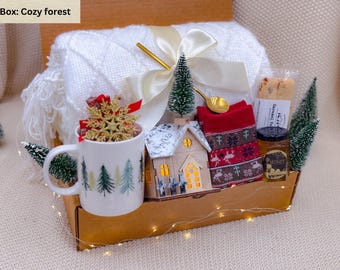May include: A cozy forest-themed gift box filled with a white blanket, a mug with a forest design, a decorative house, red socks, honey, and cookies. The box is decorated with a gold spoon, a snowflake ornament, and small Christmas trees.