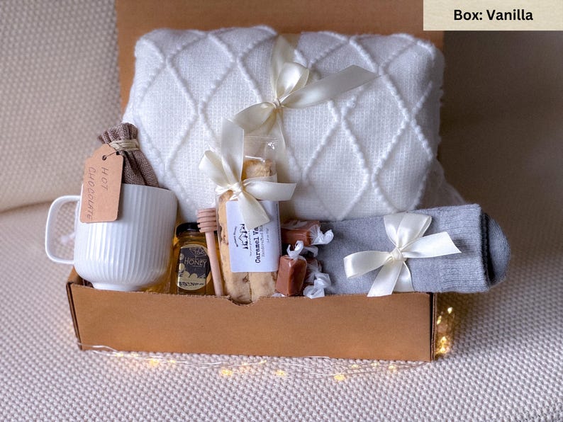 May include: A gift box filled with cozy items, including a white blanket, a mug that says "Hot Chocolate", a jar of honey, a package of biscotti, and a pair of gray socks. The box is labeled "Box: Vanilla".