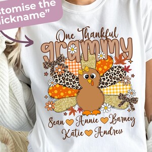 May include: White t-shirt with a Thanksgiving-themed graphic. The design features a cartoon turkey with patchwork wings, the words "One Thankful Grammy," and names below. The design includes orange, brown, and gold colours.