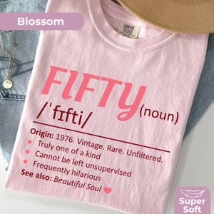 May include: A pale pink t-shirt with the word "FIFTY" in large pink letters. Below the word "FIFTY" is the phonetic pronunciation and the word "(noun)". The shirt also has a list of characteristics and the words "Super Soft".