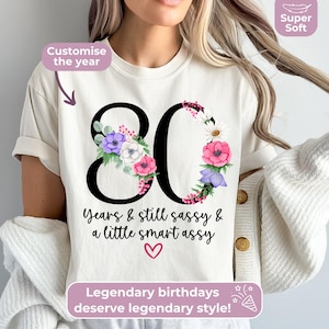 May include: A cream-colored t-shirt featuring the large black number 80 decorated with colorful purple, pink, and white floral illustrations. Below the number, the text reads Years & still sassy & a little smart assy with a small red heart.