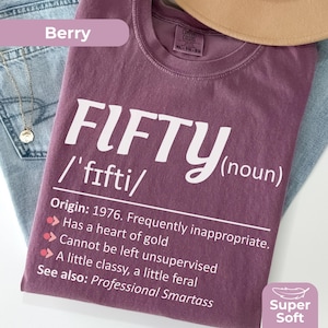 May include: Berry-coloured, XL-sized t-shirt with the word "FIFTY" in large white letters. The shirt includes a definition, origin year, and humorous characteristics. The shirt is labelled "Super Soft."