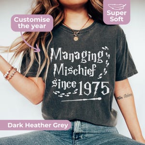 May include: A dark heather grey t-shirt featuring white text that reads Managing Mischief Since 1975, accompanied by graphic elements including lightning bolts, footprints, and a wand. The shirt includes labels for Customise the year and Super Soft.