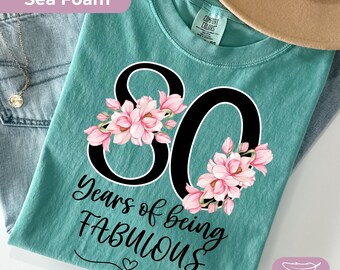 May include: Sea foam green t-shirt featuring the number 80 in black, decorated with pink floral details. The words "Years of being FABULOUS" are printed below. A tan hat and denim jeans are visible in the background.