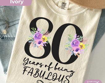 May include: An ivory t-shirt featuring a large black number 80 decorated with purple and yellow floral illustrations. Below the number, the text reads "Years of being FABULOUS" in black script and print, followed by a small black line drawing of a bow.