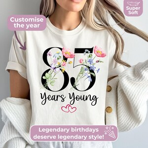 May include: A cream-coloured t-shirt featuring the large black number 85 decorated with colourful watercolour-style wildflowers. Below the number, the text reads Years Young with two small pink hearts. Text overlays include Customize the year, Super Soft, and Legendary birthdays deserve legendary style.