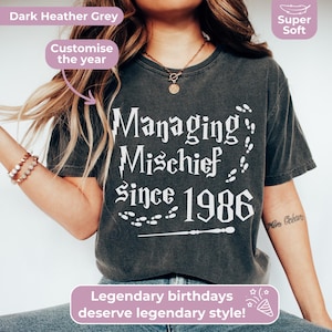 May include: Dark heather grey t-shirt with the text "Managing Mischief Since 1986" in white. The shirt also features a wand and footprints design. The text "Legendary birthdays deserve legendary style!" is also visible.