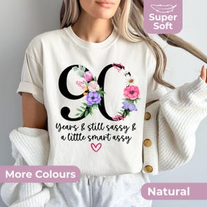 May include: A natural-coloured t-shirt featuring a large black number 90 decorated with colourful floral illustrations. Below the number, the text reads: 90 Years & still sassy & a little smart assy, accompanied by a small pink heart graphic.