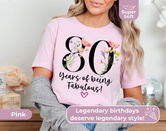 May include: A light pink short-sleeve t-shirt featuring the large black number 80 decorated with colourful floral illustrations. Below the number, the text reads "Years of being Fabulous!" in black script, followed by a small pink heart.