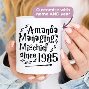 May include: White ceramic mug with black text that reads "Amanda Managing Mischief Since 1985" with a wand and footprints. The mug is being held by a person.