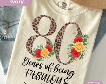 May include: An ivory t-shirt featuring a large number 80 with a brown leopard print pattern. The number is decorated with red and yellow roses and green leaves. Below the number, the text reads "Years of being FABULOUS" in black script.