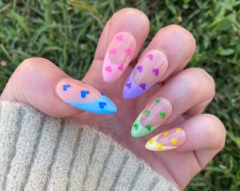 Puede incluir: Primer plano de una mano con cinco uñas en forma de almendra pintadas en diferentes colores pastel. Cada uña tiene un diseño de pequeños corazones blancos.