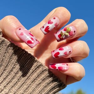 Könnte beinhalten: Nahaufnahme einer Hand mit langen, quadratischen Nägeln, die mit einem rosa-weißen Ombre-Effekt lackiert sind. Die Nägel sind mit kleinen roten Herzen und Glitzer verziert. Ein Nagel hat einen goldenen Rosen-Anhänger.