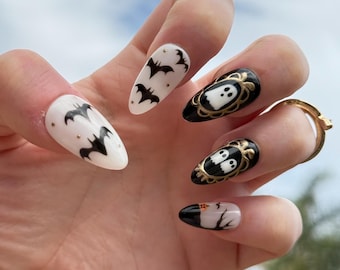 May include: Almond-shaped Halloween-themed nail art. The nails feature designs of bats, ghosts, and tree branches. The color palette includes white, black, and gold. The nails are decorated with intricate details, perfect for a spooky season look.