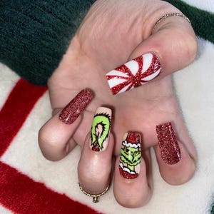 May include: A hand displaying long, square-shaped nails with Christmas-themed designs. The nail art features red glitter, a peppermint candy pattern, and Grinch character art. The nails are painted in red, green, white, and nude colours.