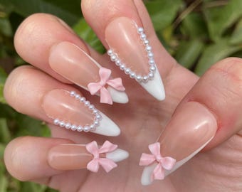 May include: Close-up of a hand with long, almond-shaped nails. The nails are painted with a white tip French manicure and decorated with pink bows and pearl accents.