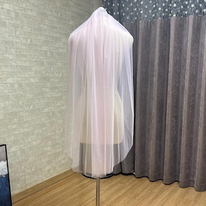 May include: A sheer, light pink bridal veil is draped over a mannequin. The veil is made of a delicate, flowing fabric. The mannequin is on a metal stand with a black base. The background includes a brick wall, curtains, and a small table.