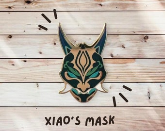 Op de afbeelding: Een gedetailleerd anime-masker van goud met groene en blauwe accenten. Het masker heeft een scherpe, puntige vorm met een woeste uitdrukking. De tekst "XIAO'S MASK" staat onder het masker.