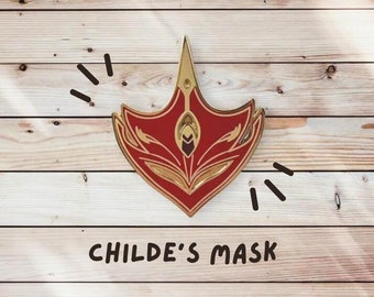 Op de afbeelding: Een rode en gouden metalen pin in de vorm van een masker met ingewikkelde details. Het masker heeft een puntige bovenkant en een gebogen onderkant. De tekst "CHILDE'S MASK" staat onder de pin.